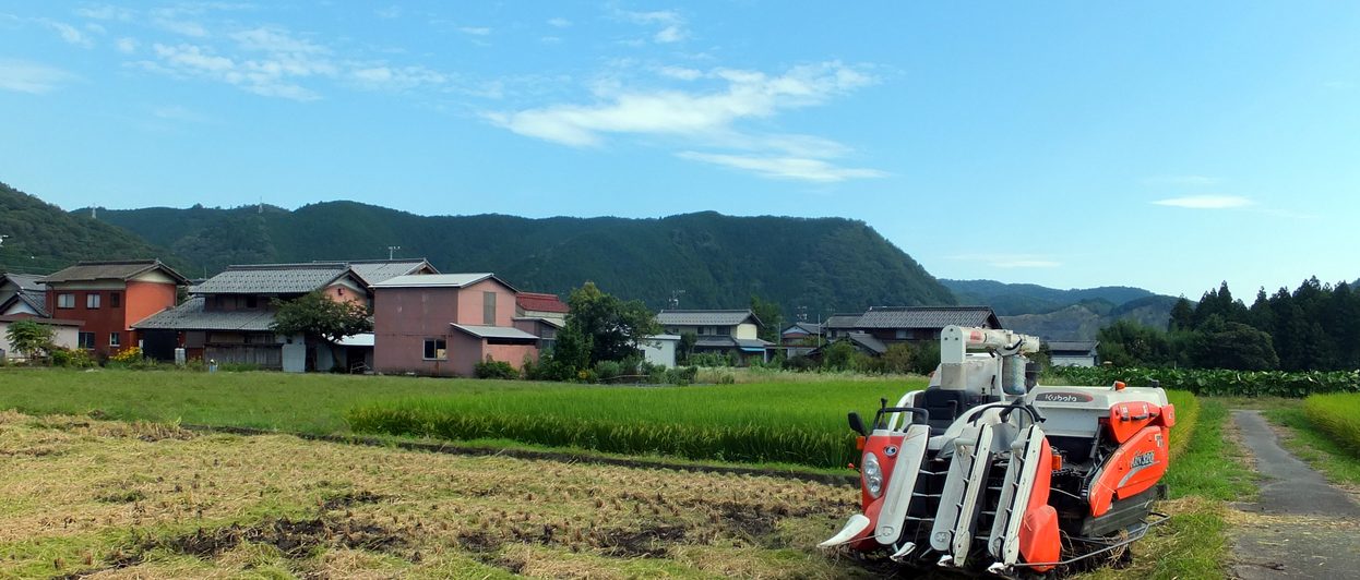 山と集落を背に、稲刈りの田んぼに止まるコンバイン
